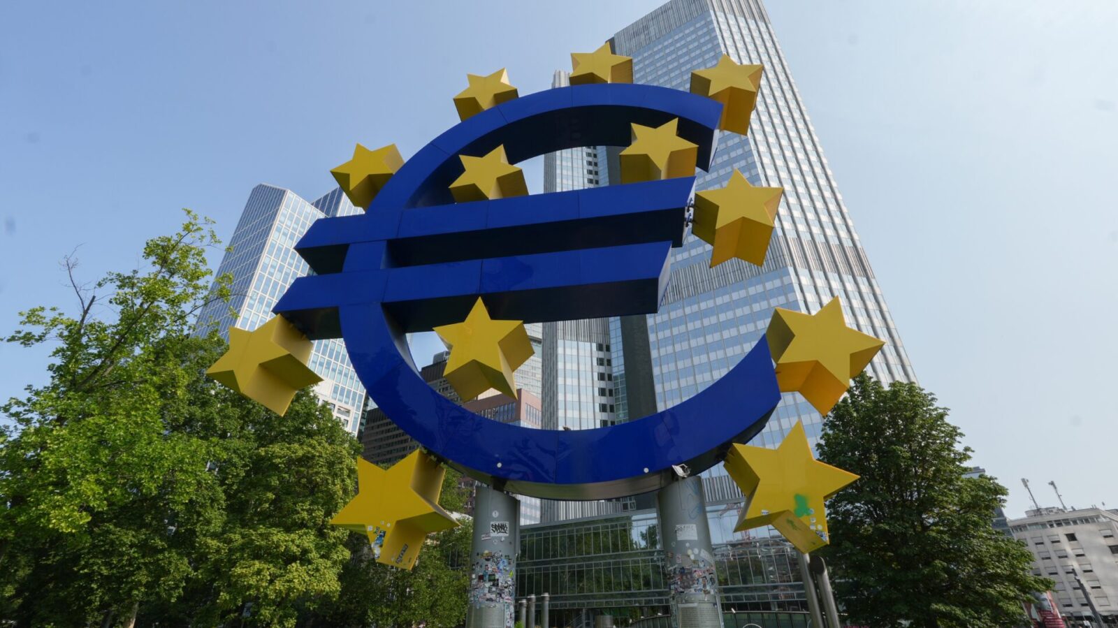 The blue and yellow euro symbol sculpture standing outside the European Central Bank in Frankfurt, with the bank's tower behind it.